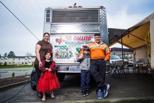 View of the back of a taco truck with a sign on it that reads "El Gallito" Mexican Food Truck. A mother and young daughter stand on the left side and a father and young son stand on the right.  