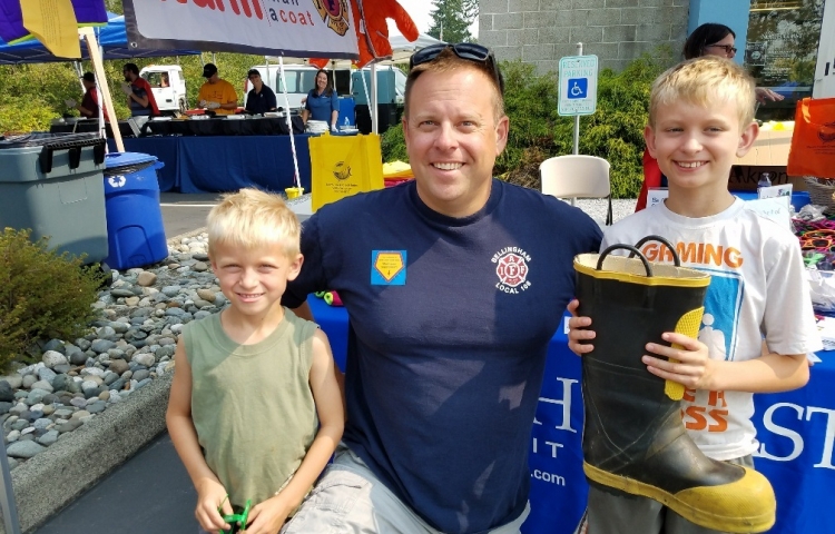 Firefighter in plain clothes kneeling with a young boy on each side of him in a outdoor festival-like setting. One of the boys is holding a firefighter's boot to collect donations for operation warm.