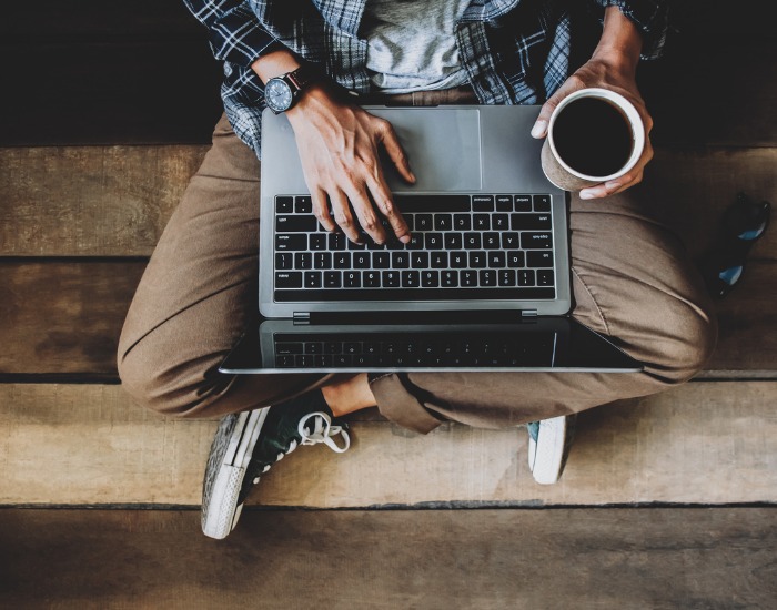 Looking down onto a man's lap. He is dressed causally and sitting cross-legged with a laptop on his lap and a mug of coffee in his hand.