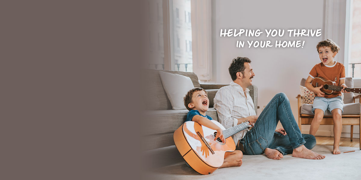A father and his two young sons are in a living room. One boy is sitting in a chair playing a ukulele and singing. The other boy is sitting in front of a couch and playing a guitar and singing next to his smiling dad. Text over the image reads helping you thrive in your home!