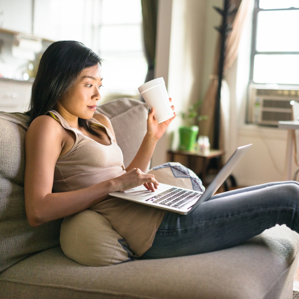 Woman lounging on a couch with a cup of coffee in her hand and working on a laptop on her lap.
