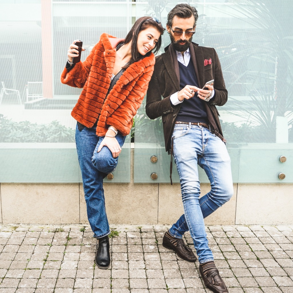 Stylish man and woman leaning up against a wall looking at the man's phone.