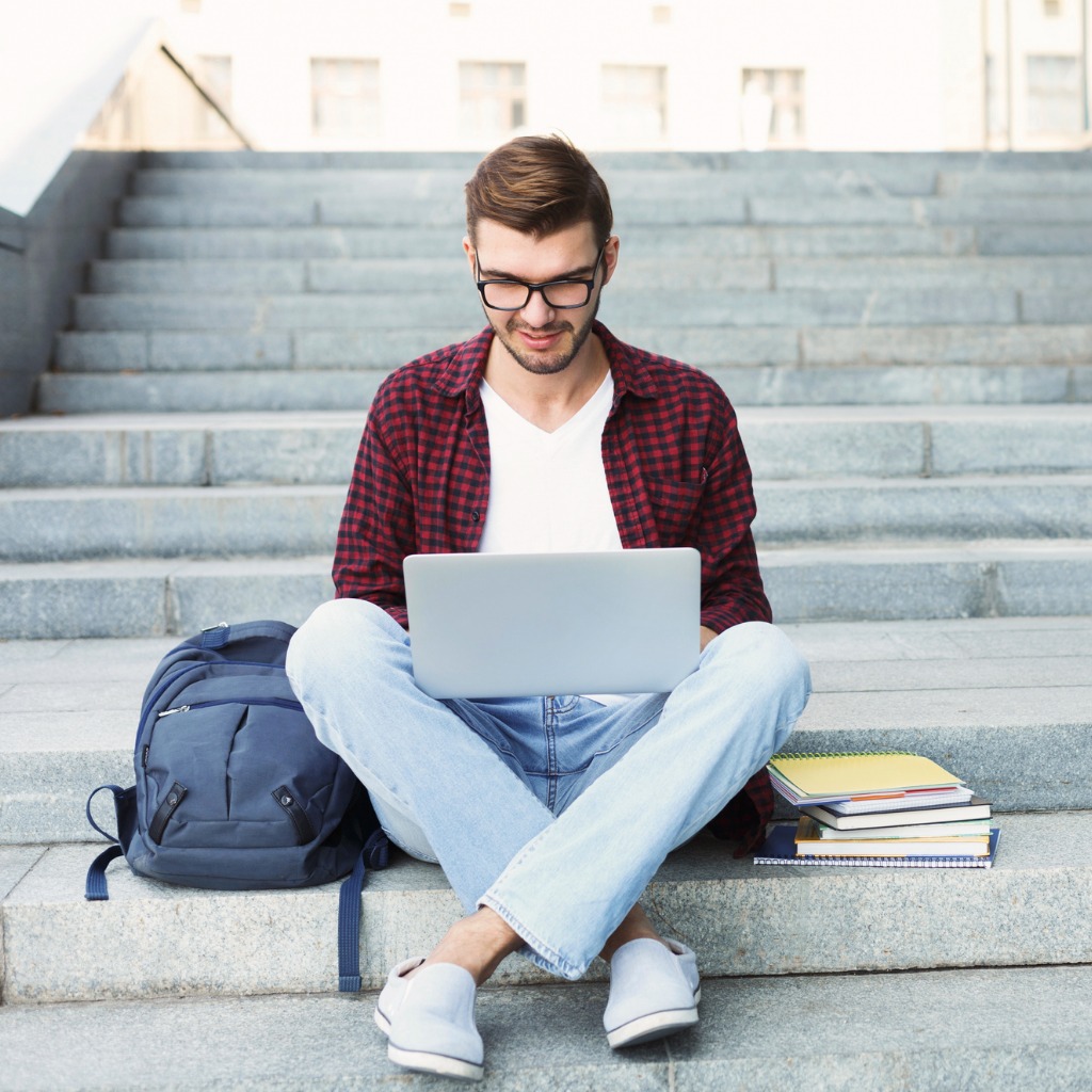 College student work on his laptop while sitting on stairs. His backpack and a stack of books surrounds him.