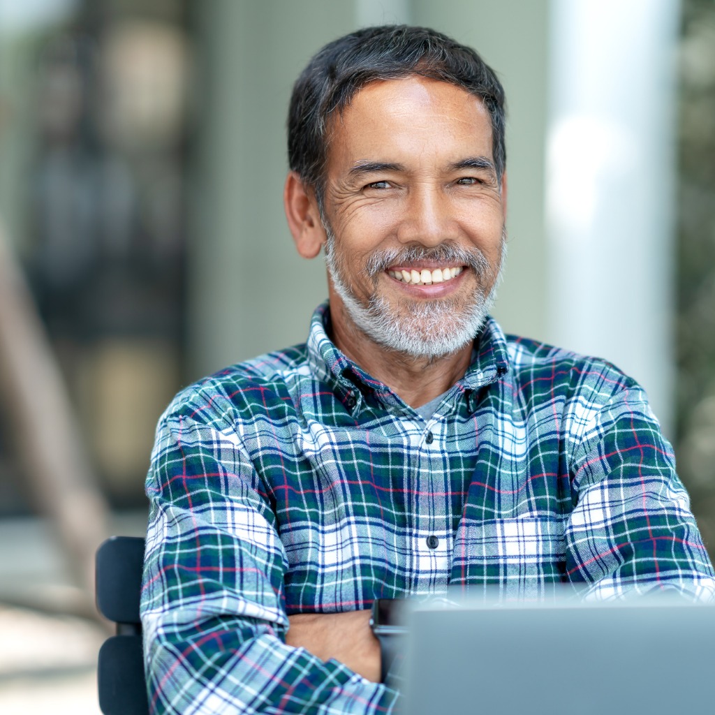 Man in blue plaid shirt with dark hair and a gray beard sits in front of a laptop with his arms folder, smiling at the camera.